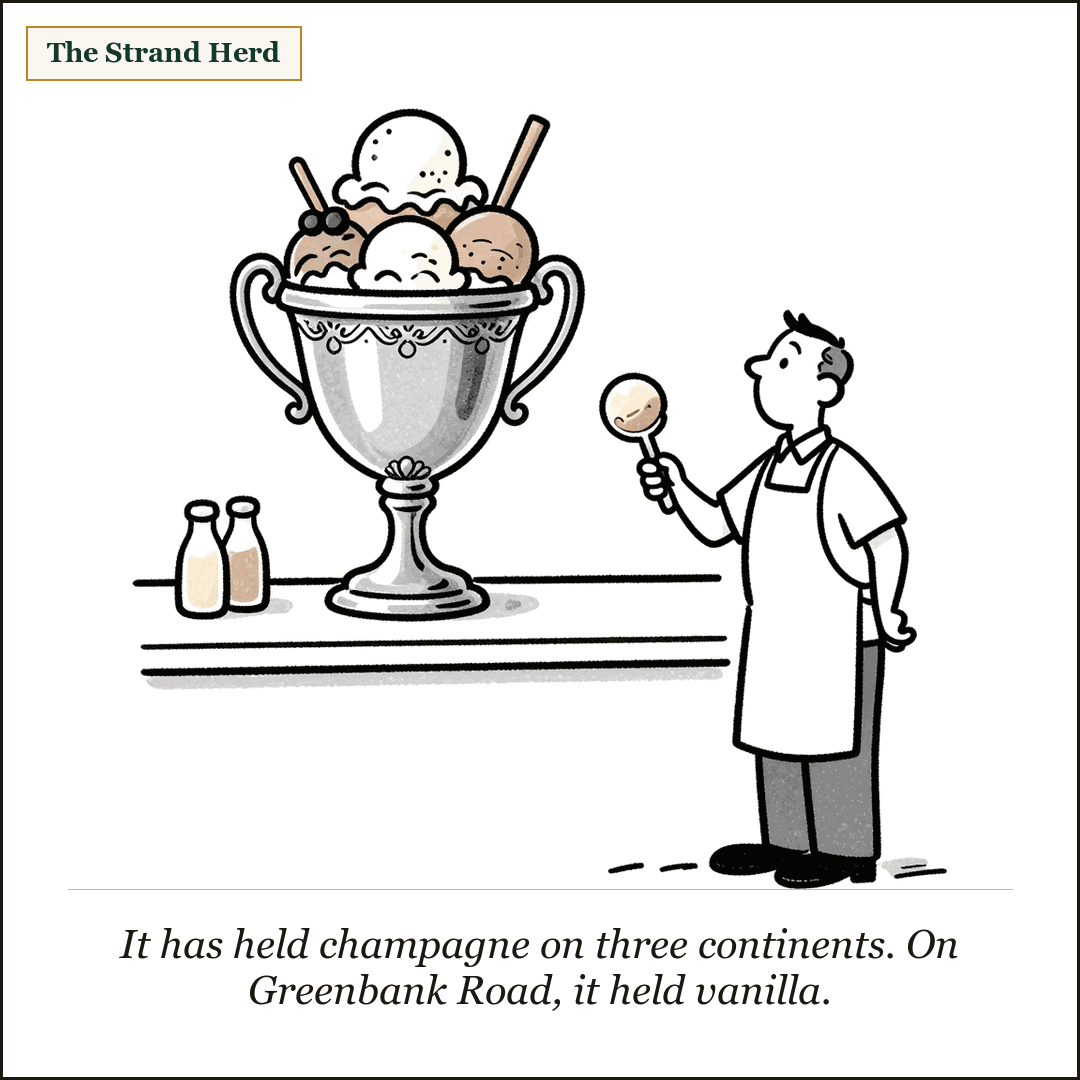 A large two-handled silver trophy on a dessert-shop counter, heaped with scoops of ice cream, while an aproned server stands beside it holding an ice-cream scoop and looking up at it.