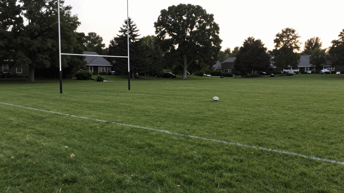An empty grass rugby pitch at dusk with white goal posts on the left and a rugby ball resting on the field, houses and parked cars visible in the background.