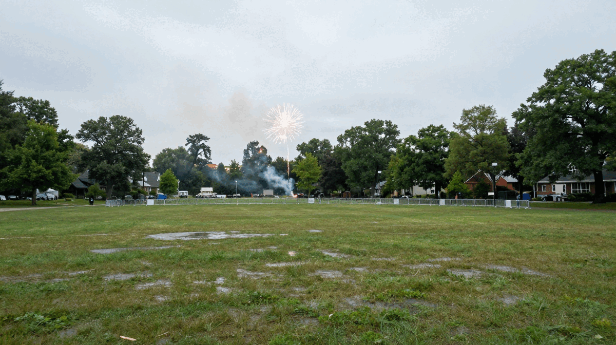 A single firework bursts in an overcast sky above a rain-soaked community park field lined with trees and houses, with smoke drifting from an earlier launch.