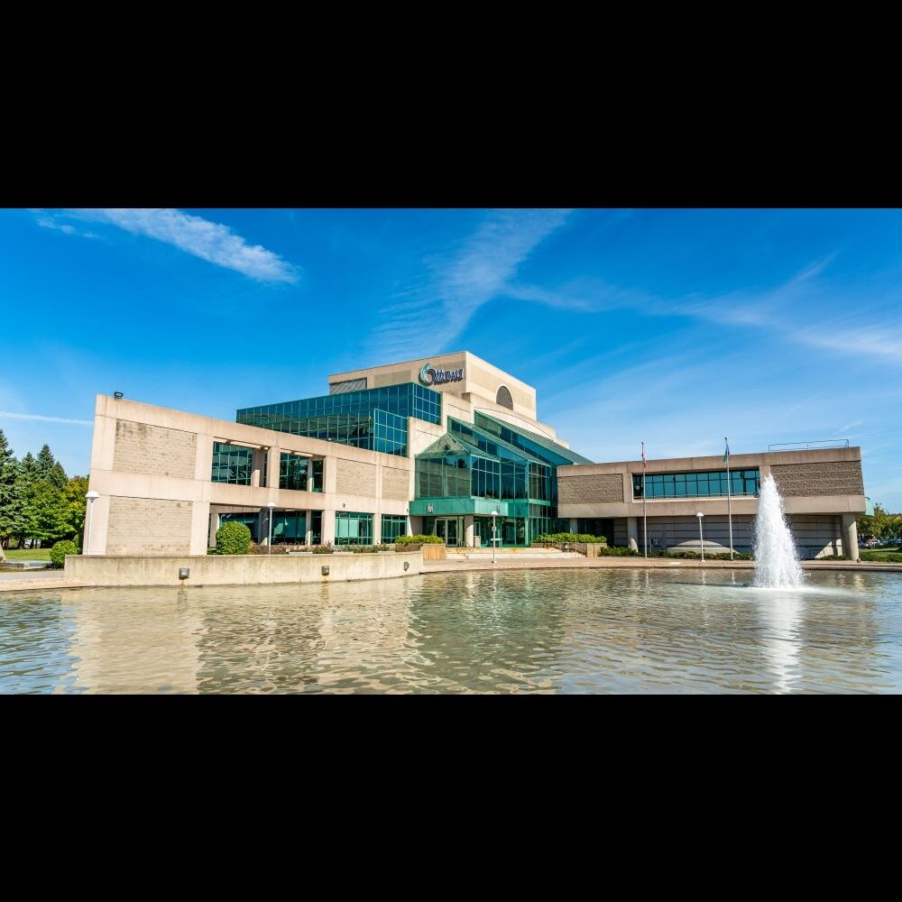 Exterior of Ben Franklin Place, a City of Ottawa municipal building with a glass atrium entrance, reflected in its fountain pond under a blue sky.