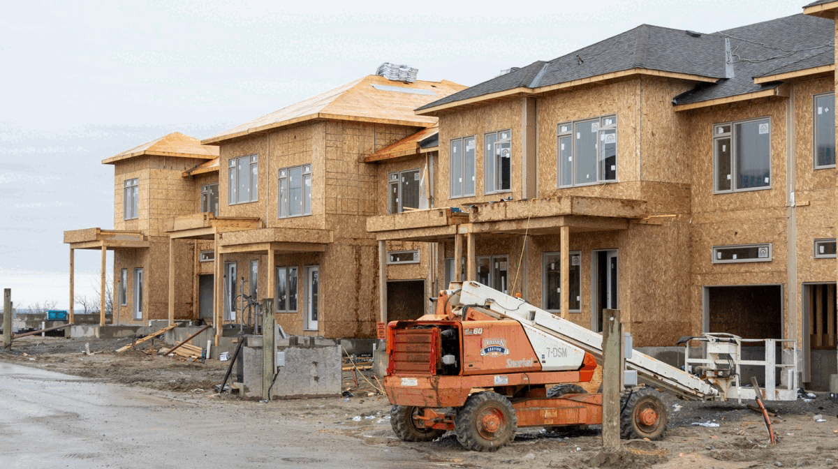 Rows of townhouses under construction in a new Ontario subdivision, framed and sheathed with plywood, roofs partly shingled, a lift parked on the muddy road in front of them.